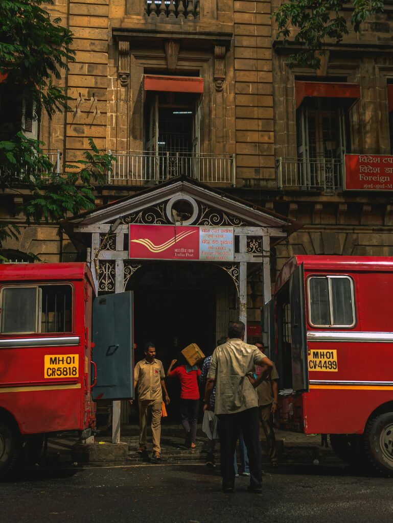 Men working at a New Delhi post office with iconic red trucks and colonial architecture.